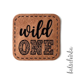 Puede incluir: Un parche de cuero marrón con un borde cosido. El texto "wild one" está impreso en negro con un patrón de leopardo.