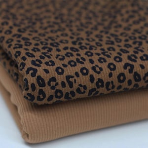 Op de afbeelding: Twee stofstalen. De bovenste stof is een bruin ribfluweel met een zwarte luipaardprint. De onderste stof is een effen bruin geribd breisel. De stoffen zijn over elkaar gevouwen.