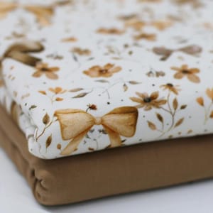 Może przedstawiać: Dwie warstwy materiału na sobie. Górna warstwa jest biała ze wzorem brązowych kwiatów i kokardek. Dolna warstwa jest w jednolitym brązowym kolorze. Materiał jest złożony, co sugeruje miękkość.