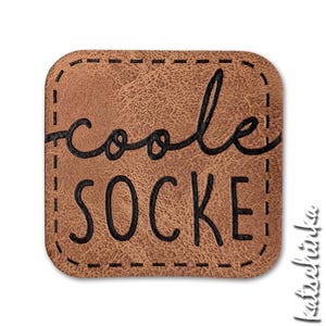 Könnte beinhalten: Ein quadratischer Aufnäher aus braunem Kunstleder mit abgerundeten Ecken und schwarzen Ziernähten am Rand. Der Text "coole" steht in schwarzer Schreibschrift über dem Wort "SOCKE" in einer fetten, schwarzen, serifenlosen Schrift auf der Oberfläche.