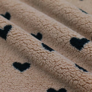 Könnte beinhalten: Nahaufnahme eines beigefarbenen Fleece-Stoffs mit einem Muster aus dunkelmarineblauen Herzen. Der Stoff ist weich und strukturiert, mit sichtbaren Falten und Schatten. Die Herzen sind gleichmäßig verteilt.
