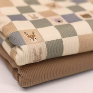 Könnte beinhalten: Ein gefalteter Rippenstoff mit einem Karomuster in Beige-, Salbeigrün- und Grautönen. Die Quadrate sind mit kleinen, einfachen Illustrationen von Waldtieren wie einem Hirsch, einem Hasen und einem Koala verziert. Ein einfarbiger brauner Stoff.