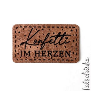 Könnte beinhalten: Ein braunes Leder-Patch mit dem Text "Konfetti im Herzen" in schwarzer Schrift.