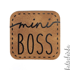 Könnte beinhalten: Ein braunes Leder-Patch mit einem genähten Rand. Der Text "mini boss" ist in Schwarz gedruckt.