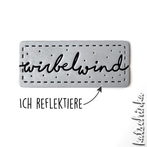 Könnte beinhalten: Ein rechteckiges silbernes Etikett mit einem genähten Rand und dem Wort "wirbelwind" in schwarzer Kursivschrift.