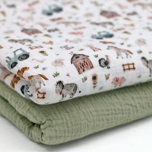 Op de afbeelding: Een witte muslin-wikkeldeken met een boerderijdierenprint. De deken heeft verschillende dieren, waaronder koeien, varkens, kippen en tractoren. De deken is dubbelgevouwen en ligt op een groene muslin-deken.