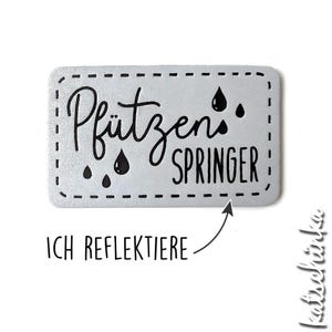 Könnte beinhalten: Ein silberner reflektierender Aufkleber mit schwarzem Text, der "Pfützen Springer" lautet, und drei schwarzen Regentropfen. Der Aufkleber hat einen gestrichelten Rand.