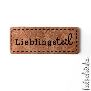Könnte beinhalten: Ein braunes Leder-Etikett mit einem genähten Rand und dem Text "Lieblingsstil" in Schwarz.