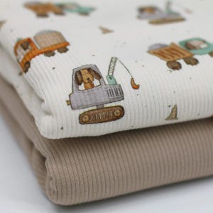 Könnte beinhalten: Zwei gefaltete Stoffe. Der obere Stoff ist cremefarben mit gerippter Textur und einem Aufdruck von Zeichentrick-LKWs und einem Hund in einem Baufahrzeug. Der untere Stoff ist einfarbig beige mit gerippter Textur.