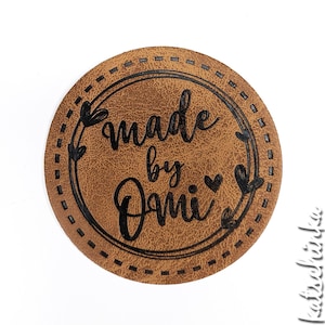 Puede incluir: Una etiqueta de cuero marrón con un borde cosido y el texto "made by Dani" en letras negras. La etiqueta presenta un diseño decorativo de corazones y hojas.