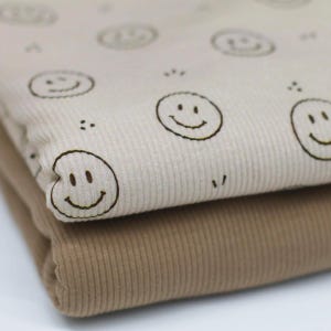 Könnte beinhalten: Zwei gefaltete Stücke aus geripptem Stoff, die übereinander liegen. Die obere Lage ist hellbeige und mit einem sich wiederholenden Muster aus dunkelbraunen, umrissenen Smileys bedruckt. Die untere Lage ist einfarbig mittelbraun und gerippt.