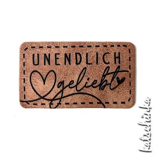 Könnte beinhalten: Braunes Leder-Patch mit einem genähten Rand. Das Patch hat schwarzen Text, der "UNENDLICH geliebt" lautet, mit einem Herzsymbol auf jeder Seite des Textes.