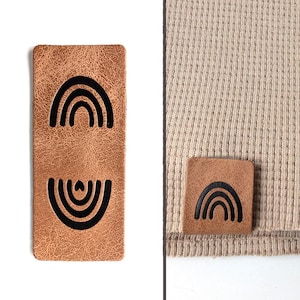 Könnte beinhalten: Zwei braune Lederpatches mit schwarzen Regenbogen-Designs. Die Patches sind auf weißem Hintergrund und auf beigefarbenem Stoff zu sehen.