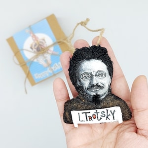 May include: A black and white felt ornament of Leon Trotsky with a black beard and a gold background. The ornament has a white banner with the text "L. Trotsky" in red.