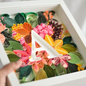 Può includere: Un'immagine incorniciata bianca con un arrangiamento di fiori di carta in tonalità di rosa, arancio, giallo e verde. La lettera "A" è ritagliata dal centro dell'arrangiamento.