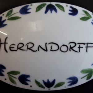 Könnte beinhalten: Ein weißes ovales Schild aus Keramik mit blauem und grünem Blumenrand und dem Wort "Herrndorf" in Schwarz.