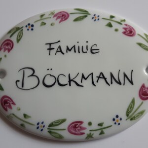 Puede incluir: Placa de puerta ovalada de cerámica blanca con un borde floral y el texto "Familie Böckmann" en negro.