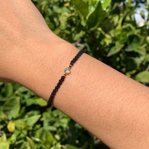 Pode incluir: Um bracelete de corda preta com uma pequena gema transparente no centro. A gema é engastada em ouro.