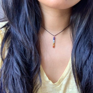 May include: A black cord necklace with a pendant made of seven small, round, colorful gemstones. The gemstones are arranged in a vertical line and are red, orange, yellow, green, blue, indigo, and violet.