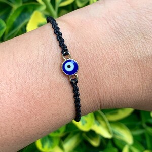 May include: Black woven bracelet featuring a gold-toned charm with a blue and white evil eye design. The bracelet is worn on a wrist, with green foliage in the background. The bracelet is a delicate piece of jewelry.