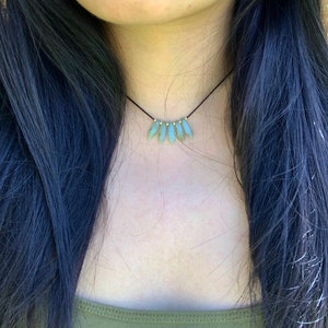 May include: A delicate choker necklace featuring a row of five light green gemstone beads, each with a gold accent. The necklace is on a black cord and is worn by a person with long black hair.