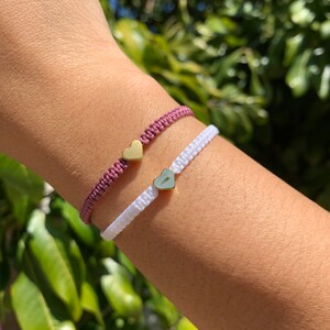 Puede incluir: Dos pulseras de la amistad con dijes de corazón de oro. Una pulsera es de color burdeos y la otra es blanca.