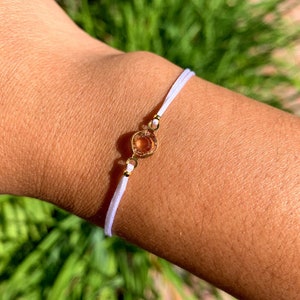 Puede incluir: Un brazalete de cuerda blanca con un dije dorado que presenta una piedra transparente de color marrón anaranjado.