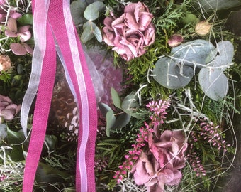 Könnte beinhalten: Nahaufnahme eines dekorativen Kranzes mit einer Mischung aus Grün, Trockenblumen und rosa Blütenzweigen. Der Kranz ist mit einem bordeauxfarbenen Band und einem durchsichtigen weißen Band verziert, was eine natürliche und elegante Ästhetik schafft.