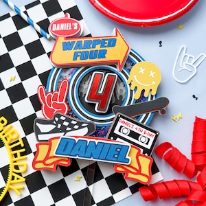 May include: A colorful birthday cake topper with the name "Daniel" and the words "Warped Four". The design includes a skateboard, a cassette tape, a shoe, and a smiley face. A red plate and a checkered flag are also visible.