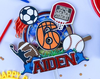 May include: A sports-themed cake topper with a blue, red, and white color scheme. It features a basketball, baseball, soccer ball, and a football. The topper also includes the words "It's Game Time!" and "Aiden".