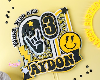May include: A yellow, black, and white birthday cake topper with the name "AYDON" and the number "3". The topper includes a peace sign, a smiley face, and the words "YOUNG, WILD AND". The topper is on a clear stick.