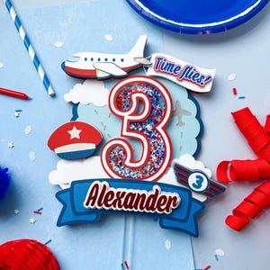 May include: A blue, red, and white airplane-themed cake topper with the number 3, the name "Alexander", and the phrase "Time flies!". The topper includes a small airplane, clouds, and a pilot's hat. The background is light blue.