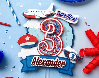 May include: A blue, red, and white airplane-themed cake topper with the number 3, the name "Alexander", and the phrase "Time flies!". The topper includes a small airplane, clouds, and a pilot's hat. The background is light blue.