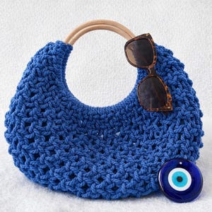 May include: A vibrant blue macrame handbag featuring a crescent shape and a rounded light wood handle. Resting on the bag are brown tortoiseshell sunglasses, and a circular blue and white glass evil eye charm is positioned at the bottom right.