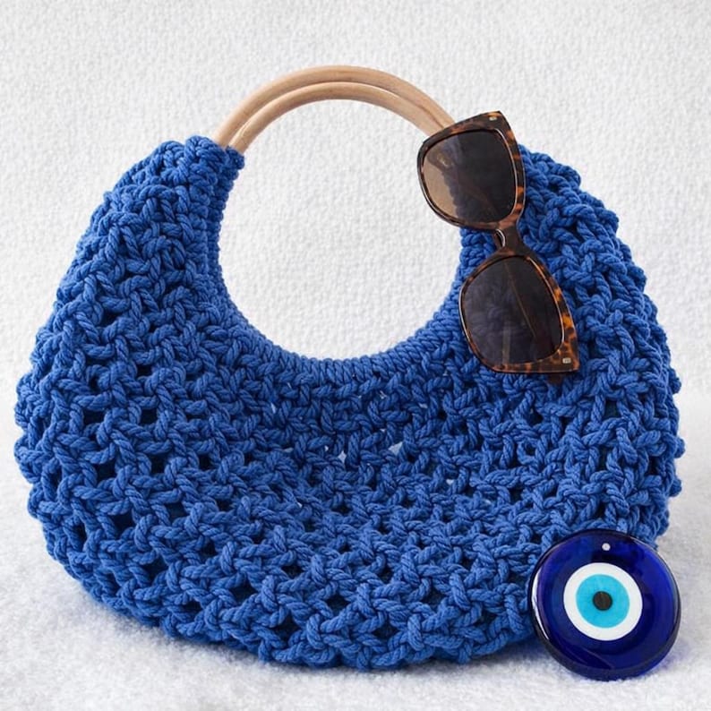 May include: A vibrant blue macrame handbag featuring a crescent shape and a rounded light wood handle. Resting on the bag are brown tortoiseshell sunglasses, and a circular blue and white glass evil eye charm is positioned at the bottom right.