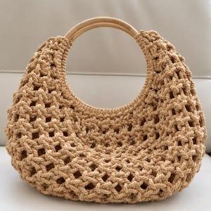 May include: A beige macrame handbag featuring a crescent-shaped body with an open-weave knotted pattern and a smooth, circular light-colored wooden handle integrated into the top opening of the bag structure.