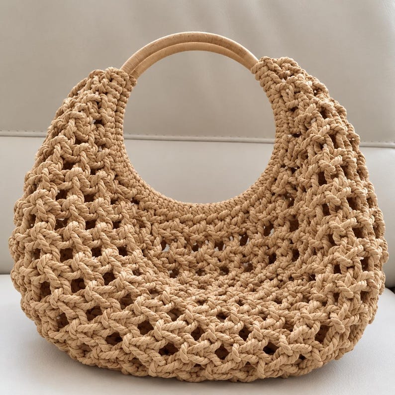 May include: A beige macrame handbag featuring a crescent-shaped body with an open-weave knotted pattern and a smooth, circular light-colored wooden handle integrated into the top opening of the bag structure.