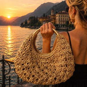 May include: A gold-toned, crocheted handbag featuring a crescent-shaped silhouette and a large, smooth, circular metallic handle. The bag is constructed with an open-weave, knotted pattern that creates a textured, shimmering appearance throughout.