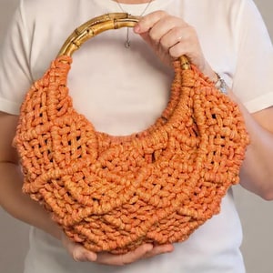 May include: A bright orange macrame handbag featuring a textured, knotted cord pattern and a curved, natural bamboo handle. The bag has a crescent-shaped silhouette with an intricate woven design throughout the body, providing a structured yet flexible appearance.