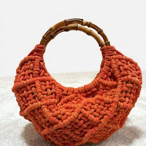 May include: A crescent-shaped handbag crafted from orange cord in a complex macrame knot pattern. The bag features two rounded, natural bamboo handles attached to the top, providing a structured contrast to the soft, textured orange body.