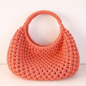 May include: A coral-colored handbag crafted with a dense, knotted macrame texture. The bag features a rounded, semi-circular silhouette and a single, integrated circular handle that is wrapped in a smooth, ribbed knit material for a uniform look.