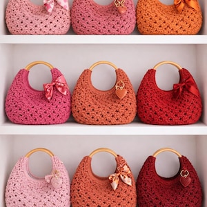 May include: Nine crescent-shaped crochet handbags arranged on three shelves. Each bag features a round wooden handle and a textured open-weave pattern in various shades of pink, orange, and red, accented with small satin bows or heart charms.