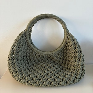 Può includere: Una borsa a mano color verde salvia realizzata con corda spessa in un motivo a nodi macramè. La borsa presenta un corpo arrotondato a forma di mezzaluna e un unico manico circolare integrato, rivestito con la stessa corda verde salvia.