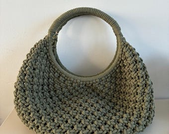 May include: A sage green handbag crafted from thick cord in a macrame knot pattern. The bag features a rounded, crescent-shaped body and a single integrated circular handle that is wrapped in the same sage green cord material as the main body.