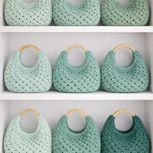May include: Nine crocheted handbags arranged on shelves, each featuring a rounded wooden handle and a textured, open-weave pattern. The bags are displayed in a gradient of green shades, ranging from pale mint to deep forest green, with a consistent crescent shape.