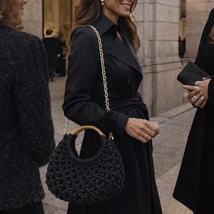 May include: A black crocheted handbag featuring a rounded silhouette, a smooth light-colored wooden circular handle, and a gold-toned metal chain shoulder strap. The bag has a textured, knotted surface pattern and is worn with a black trench coat.
