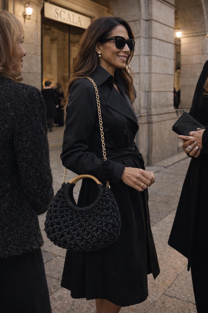 May include: A black crocheted handbag featuring a rounded silhouette, a smooth light-colored wooden circular handle, and a gold-toned metal chain shoulder strap. The bag has a textured, knotted surface pattern and is worn with a black trench coat.