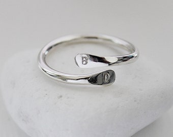 May include: A silver ring with a simple, open design. The ring is made of a single band of metal that is twisted and bent to create a unique shape. The ring is engraved with the letter "D" on one side and the letter "B" on the other.