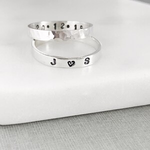 May include: Two silver rings stacked on top of each other. The top ring is hammered and has numbers engraved on it. The bottom ring is smooth and has the initials "J ♥ S" engraved on it.