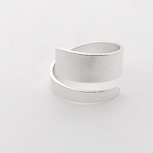 May include: A silver ring with a wide, flat band that wraps around the finger. The band is smooth and has a brushed finish.